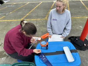 Two people crouch over a blue rectangle plastic container. One holds a white notepad, the other holds a measuring ruler. An orange magnifying glass sits on the storage container.