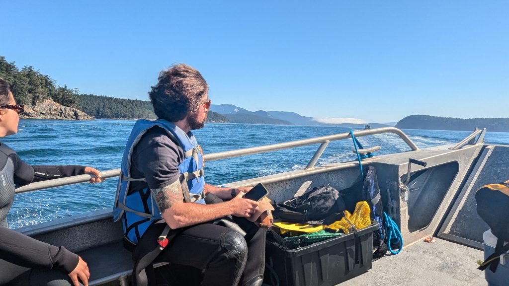 Two people sit on a boat, one wearing a blue life jacket, the other a black wetsuit.