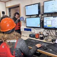 Someone wearing an orange hardhat and red life jacket, sits at a computer table looking at multiple screens with data showing.