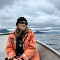 A woman wearing an orange life jacket sits on a boat on a large lake.