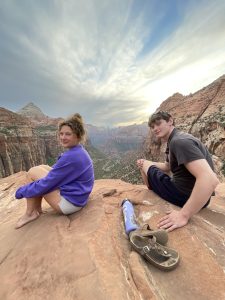 Two people sit on a cliff overlooking a valley