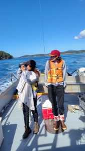 Two people stand on a boat looking to the left, one with binoculars.