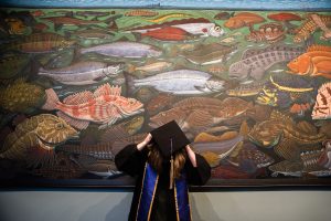 Owen Proulx puts on graduation cap in front of fish mural