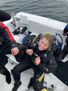 Cali Weber sits in scuba diving gear on a boat, smiling up at the camera.