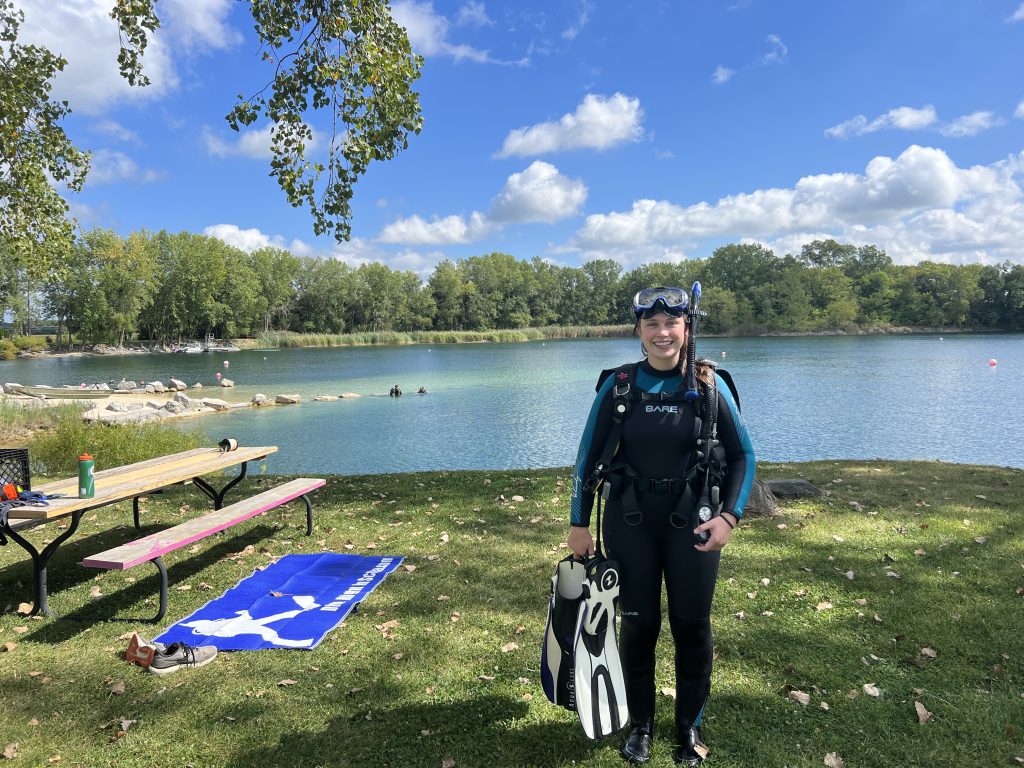 Cali Weber stands on the grass by a filled in quarry, wearing scuba diving gear.