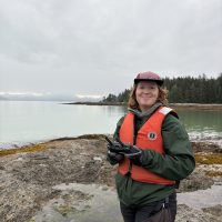 Bella Andre (ESRM/Marine Biology) stands on a rocky coastline with water in the background, holding a drone controller.