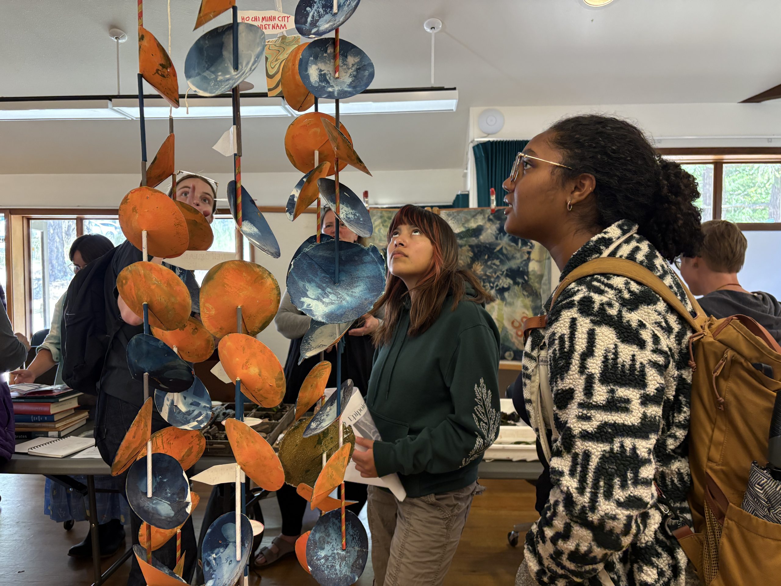 A group of people admire an art exhibit, colored orange and blue, hanging from the ceiling.